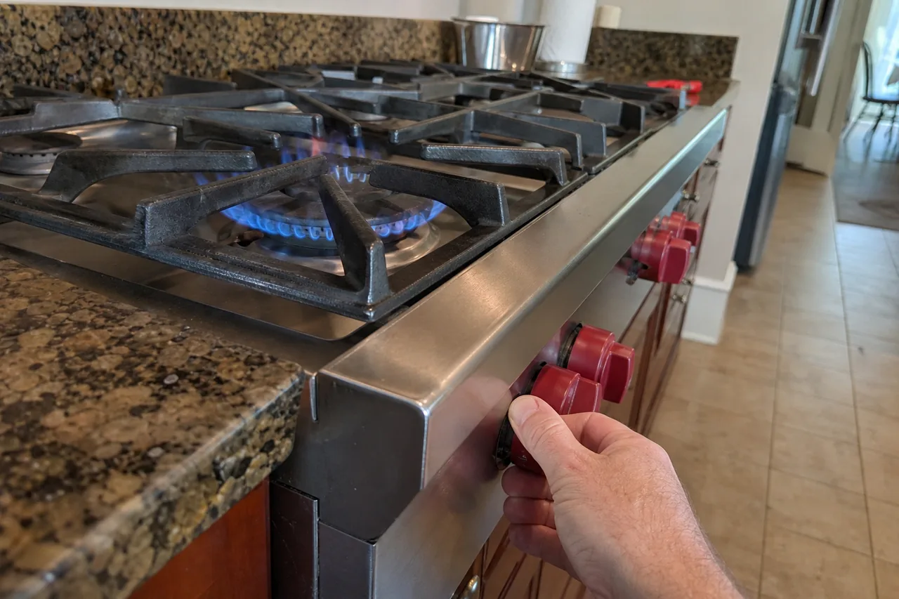 A hand turning one of the red control knobs on the front of a stainless gas rangetop, a ring of blue flame burning under a heavy cast-iron grate set into a granite counter.