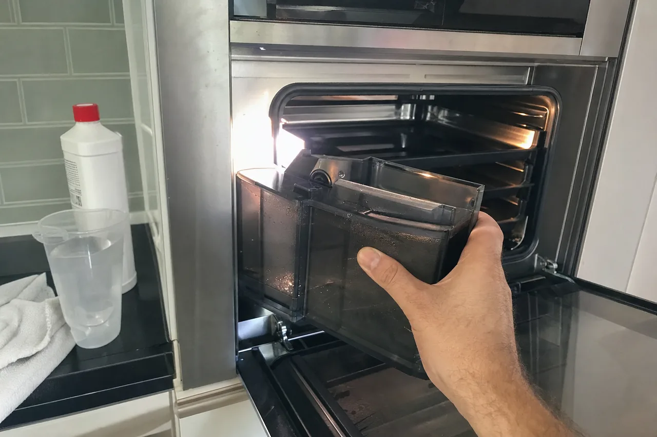 The open cavity of a built-in steam oven with its reservoir drawn out and a measuring beaker beside it