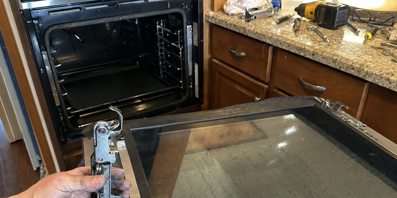 An open built-in oven cavity with its door carried clear onto the worktop, a hand under the hinge arm and a cordless drill on the granite behind